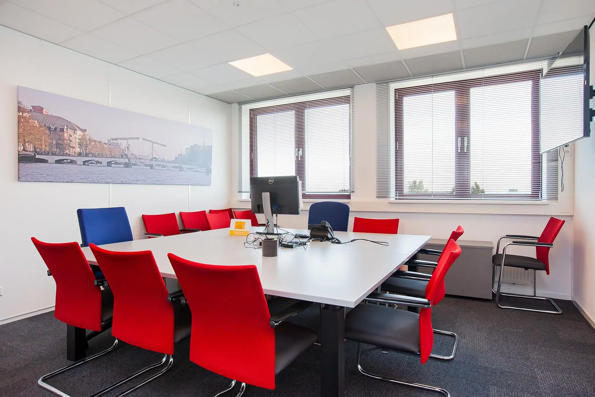 Vergaderruimte met rood-zwarte stoelen, een groot scherm en een foto van de Magere Brug in Amsterdam aan de muur.